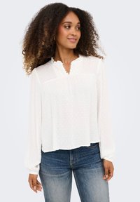 Γυναίκα με σγουρά μαλλιά, φοράει λευκό μπλουζάκι με μακριά μανίκια και υφή, καθώς και μπλε τζιν, κοιτάζοντας πλάγια με ένα ελαφρύ χαμόγελο.