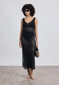 Μαύρο κροσέ φόρεμα κάλυψης με γεωμετρικό μοτίβο, χωρίς μανίκια και με σκίσιμο έως τον μηρό. Το μοντέλο κρατά μια τζιν τσάντα σε μαύρη και καφετί ρίγα.