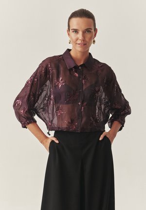 Przezroczysta, ciemnofioletowa bluzka z haftem kwiatowym z cekinów, z przodu zapinana na guziki, z szerokimi rękawami i ściąganymi mankietami, zestawiona z czarnymi spodniami o wysokim stanem.