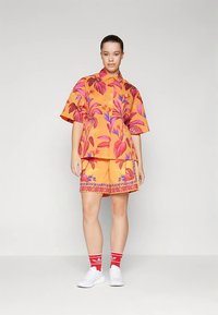 Γυναίκα στέκεται φορώντας ταιριαστό πορτοκαλί πουκάμισο και σορτς με ροζ και μωβ λουλούδια, κόκκινες κάλτσες και λευκά αθλητικά παπούτσια μπροστά σε γκρίζο φόντο.