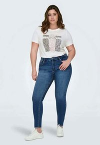 T-shirt graphique blanc avec des motifs variés et du texte, associé à un jean skinny bleu foncé. Le modèle porte des baskets blanches. Tissu lisse, design ajusté.