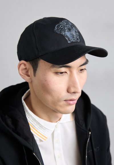 Casquette en coton noir avec un visière structurée, ornée d'un motif de méduse brodé. Portée avec un sweat à capuche noir et une chemise blanche avec des accents de col.