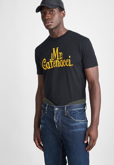 T-shirt en coton noir avec impression jaune "Mr. Catenacci", associé à un jean en denim bleu avec une taille côtelée verte. Le modèle est debout.