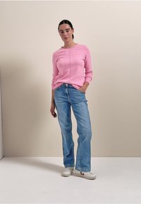 Rosa gestrickter Pullover mit Rundhalsausschnitt, kombiniert mit hellblauen, locker sitzenden Jeans und weißen Sneakers. Schlichtes, lässiges Outfit vor einem neutralen Hintergrund.