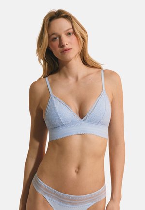 Frau trägt hellblauen Spitzen-Bralette mit verstellbaren Trägern und passende, spitzenbesetze Unterwäsche vor einfarbigem Hintergrund.