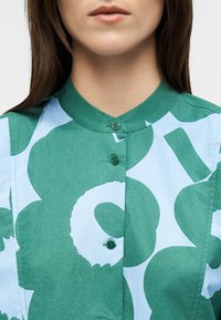 Femme portant une chemise à motifs verts et bleu clair avec un col rond et trois boutons verts visibles, seules les lèvres et le menton sont visibles, aucun autre détail n'est montré.