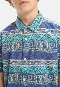 Korte mouwen overhemd met knopen, voorzien van een veelkleurig geometrisch patroon in turquoise, paars en crème. Gemaakt van lichte stof.