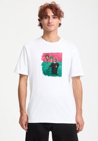Hvid kortærmet t-shirt med et farverigt grafisk motiv af en karakter med en pigget sol og et træ på en grøn baggrund.