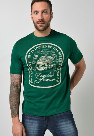 Mężczyzna z tatuażem na ramieniu, ubrany w zielony t-shirt z czaszką w bandanie i napisem "Duch Żeglarza kształtowany jest przez bezlitosne morze, niezłomny marynarz."