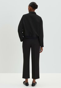 Zwarte cropped jack met kraag en geribbeld zoom, gecombineerd met zwarte wijde broek. Glad materiaal en getailleerde pasvorm zichtbaar vanaf de achterkant.