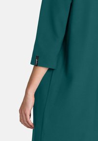 Rochie teal cu mâneci trei sferturi și un luciu subtil. Prezintă un etichetă la manseta și o textură de țesătură netedă și fluidă.