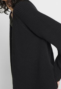 Personne portant un cardigan noir en tricot épais avec des manches amples sur un haut vert foncé, sur un fond clair uni.