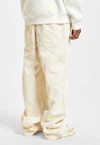 Beige Weite-Leg Cargo-Hosen aus robustem Baumwollmaterial mit sichtbaren Taschen, Nahtdetails und einem lockeren Schnitt, kombiniert mit beigen Sportschuhen.