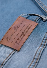 Étiquette en cuir marron avec texte en relief, attachée à un tissu denim bleu clair, présentant une bordure cousue et une boucle pour suspension.