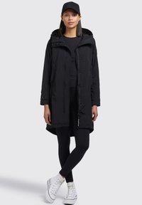 Svart vattentät parkas med huva, med dragkedja och tryckknappar, matchad med svarta leggings och vita högstövlar.