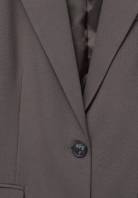 Blazer marron en tissu texturé, avec un revers cranté, une fermeture par un bouton noir unique et une doublure intérieure élégante.