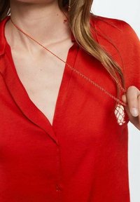 Blouse rouge transparente avec un col et des boutons. Un délicat collier en chaîne d'or avec un pendentif texturé pend du décolleté.