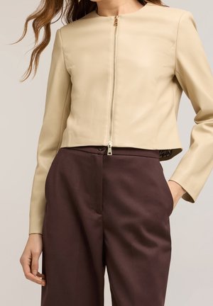 Beige kort jakke med frontlynlås kombineret med højtaljede mørkebrune bukser, model stående med den ene hånd i lommen.