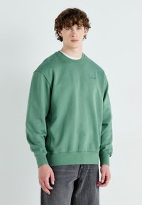 Groene katoenen sweatshirt met geribde manchetten en onderkant, voorzien van een ronde halslijn en subtiele logodetail op de borst. G gedragen met grijze broek.