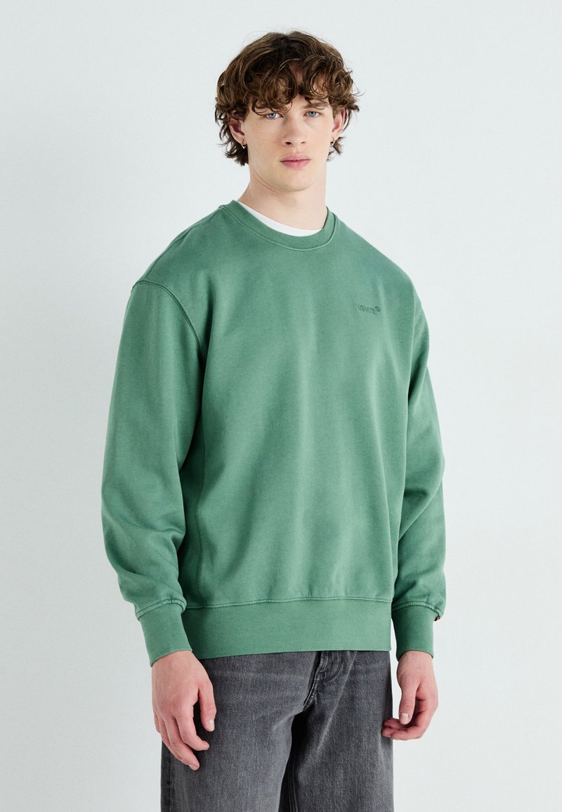 Groene katoenen sweatshirt met geribde manchetten en onderkant, voorzien van een ronde halslijn en subtiele logodetail op de borst. G gedragen met grijze broek.