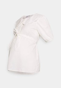 Camisa branca com colarinho em formato de ondas, mangas curtas bufantes e um cinto amarrado na frente. Feita de um tecido leve com ligeira textura.