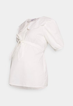 Camisa branca com colarinho em formato de ondas, mangas curtas bufantes e um cinto amarrado na frente. Feita de um tecido leve com ligeira textura.