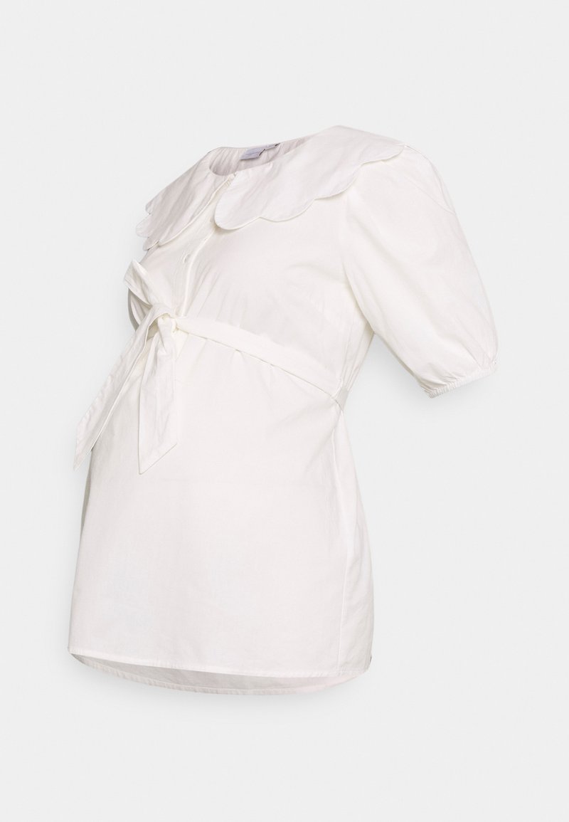 Camisa branca com colarinho em formato de ondas, mangas curtas bufantes e um cinto amarrado na frente. Feita de um tecido leve com ligeira textura.