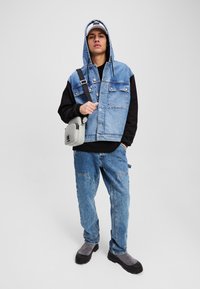 Denim vest met capuchon, zwarte mouwen en meerdere zakken, gedragen met lichtblauwe jeans en grijze enkellaarzen. Draagt een grijze schoudertas.