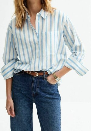 Vrouw draagt een lichtblauw-wit gestreept overhemd met verticale strepen, ingestopt in een hooggesneden donkerblauwe spijkerbroek met een bruine riem en een gouden horloge.