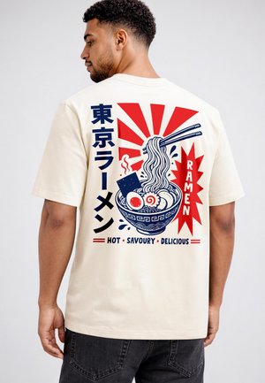 Mann trägt beiges T-Shirt mit buntem Aufdruck einer dampfenden Ramen-Schüssel, Essstäbchen, japanischem Text und englischen Worten "Ramen Hot Savoury Delicious" auf dem Rücken.