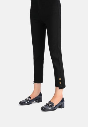 Nogi w czarnych, dopasowanych spodniach z złotymi guzikami na kostkach oraz granatowych loafersach na niskim obcasie z srebrnymi klamrami.