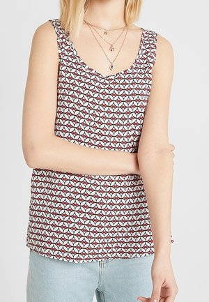 Person wearing sleeveless patterned top with red, black, and white geometric design, light blue jeans, and layered gold necklaces.