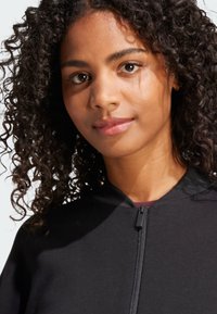Chaqueta negra con cremallera, de textura suave y cuello acanalado, con una vista detallada del rostro de un modelo y su cabello rizado.