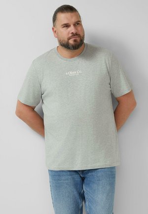 Homme à la barbe portant un t-shirt gris clair de la marque s.Oliver Co. et un jean bleu, debout les mains derrière le dos contre un mur gris uni.