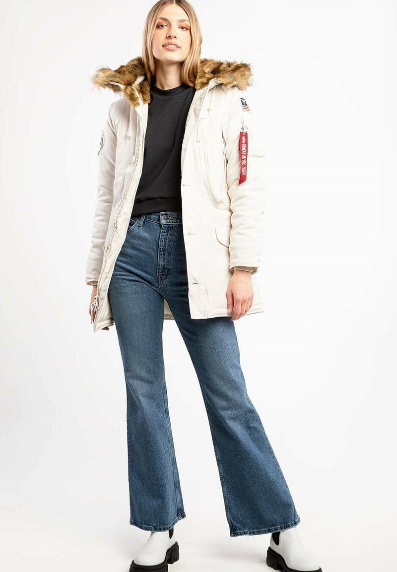 Weißer Parkajacke mit Fellkapuze, Reißverschluss und Druckknopfverschlüssen sowie seitlichen Taschen. Getragen über einem schwarzen Oberteil, kombiniert mit blauen Schlagjeans und schwarzen Stiefeln.