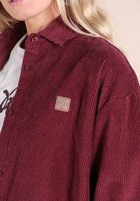 Chemise en velours côtelé bordeaux avec col, présentant une texture côtelée visible et une petite étiquette carrée sur la poitrine. Boutonnée au niveau du cou.