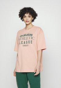 Rožinė per didelė marškinė, pagaminta iš medvilnės, su tamsiai žaliu užrašu "adidas ATHLETIC LEAGUE". Trumpomis rankovėmis ir apvaliu kaklu.
