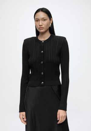 Femme aux longs cheveux noirs portant un cardigan noir cintré à boutons avec des motifs texturés et une jupe noire sur un fond clair uni.