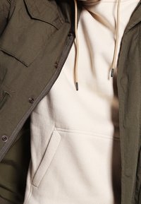 Olijfgroen jack met een textuur oppervlak, gelaagd over een romig beige hoodie met een voorvak en een capuchon met trekkoord.