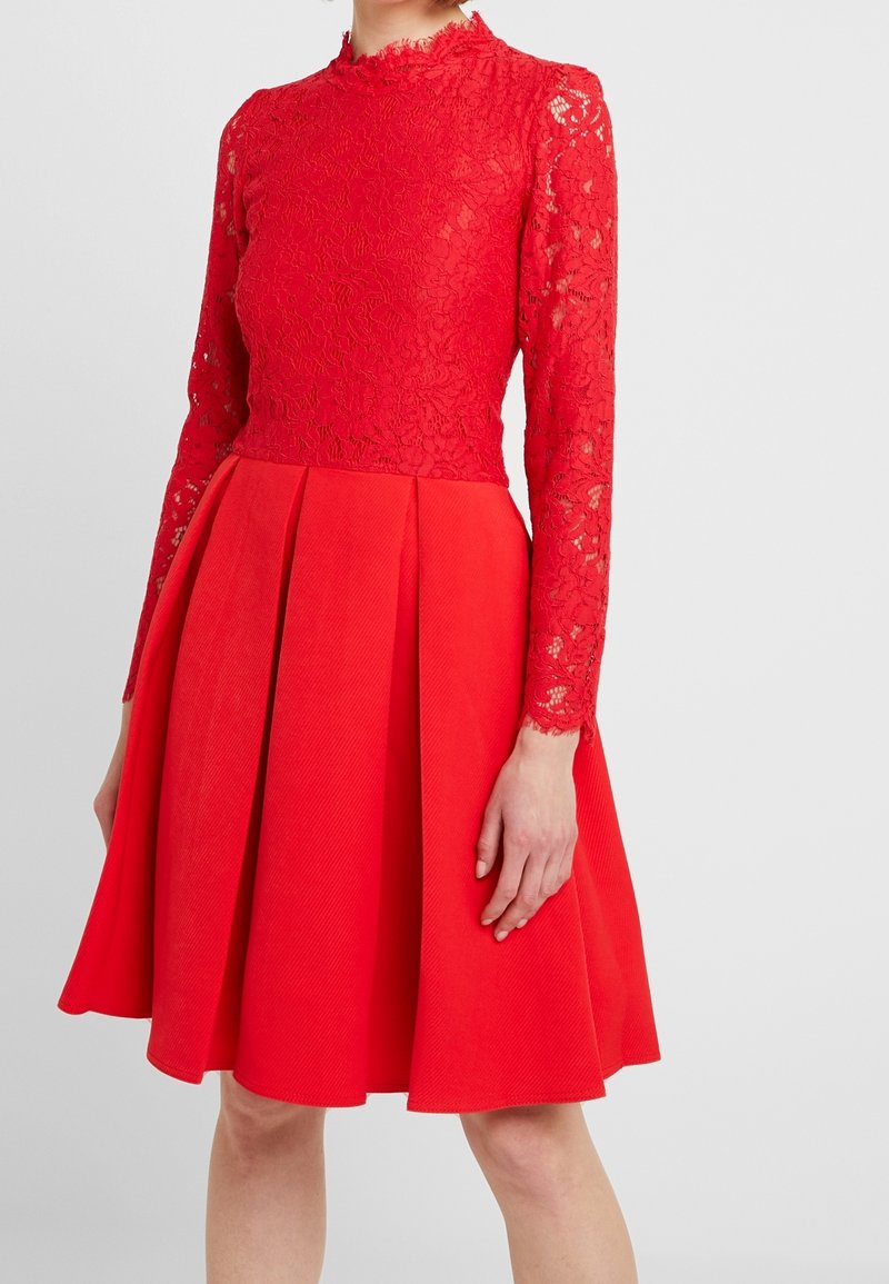 Femme portant une robe rouge longueur genou avec des manches longues en dentelle et une jupe plissée, debout devant un fond clair uni.