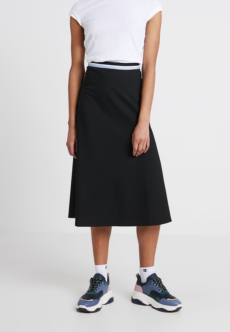 Jupe noire trapèze en tissu lisse, taille haute avec poches latérales, associée à un t-shirt blanc à manches courtes et des baskets épaisses.