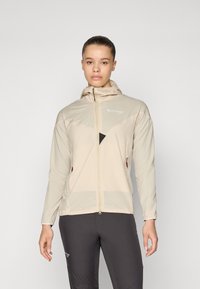 Veste légère beige avec capuche, dotée d'une fermeture éclair et d'accents en maille. Comprend des poches latérales avec des détails zippés.