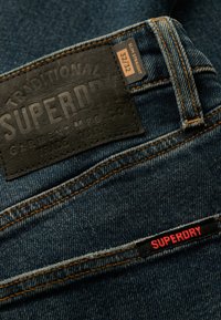 Vaqueros de mezclilla azul con una etiqueta de cuero negro texturizada grabada con "SUPERDRY". Presenta costuras naranjas y una pequeña etiqueta roja "SUPERDRY".