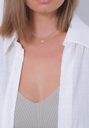 Eine medalsichelblumenförmige Halskette an einer geflochtenen Kette, die über einem hellgrauen Oberteil und einem weißen Shirt getragen wird, hebt das filigrane Design und den Glanz hervor.
