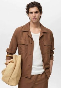 Brun knappet jakke med to brystlommer, båret over en hvid top. Modellen holder en struktureret beige taske.