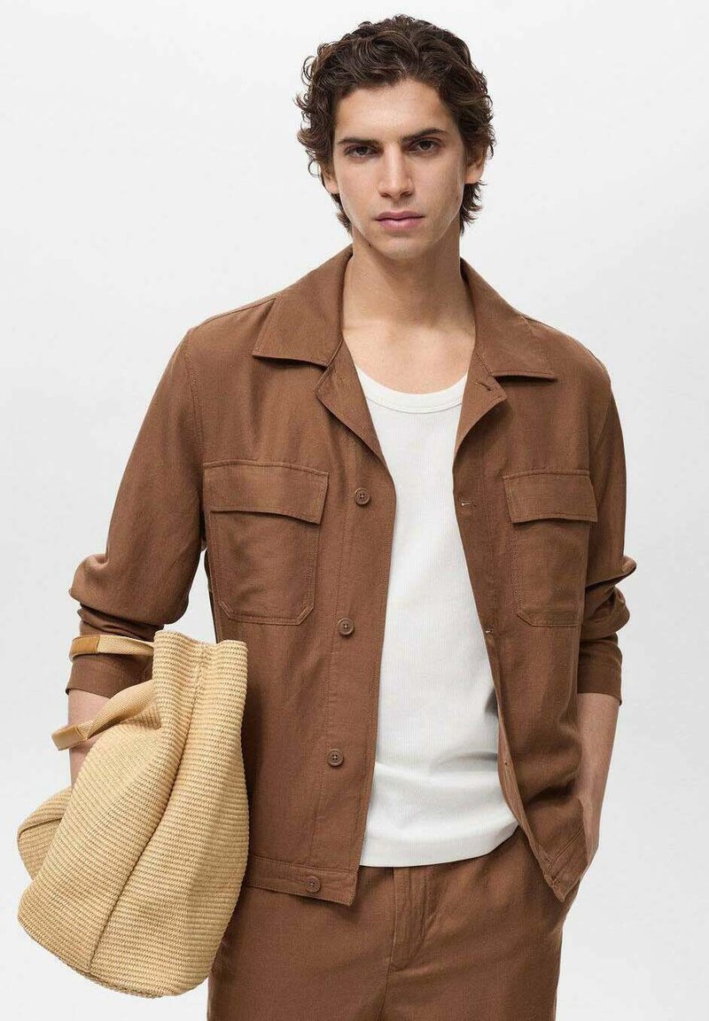 Brun knappet jakke med to brystlommer, båret over en hvid top. Modellen holder en struktureret beige taske.