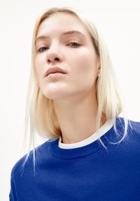 Blauwe trui met een ronde hals, textuur gebreid en witte rand. Het model heeft lang blond haar, een neutrale uitdrukking en minimale make-up.