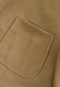 Hnědá tkanina s hladkým povrchem, která má obdélníkovou kapsu a čisté švy. Materiál vypadá měkký a odolný.