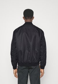 Giubbotto bomber nero con una texture liscia e lucida, colletto e polsini a costine, e maniche arricciate. La vista posteriore mostra un design e una vestibilità aerodinamici.