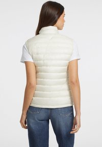 Helle cremefarbene, gepolsterte Weste mit hohem Kragen und horizontalen Steppnähten. Getragen über einem weißen Hemd und kombiniert mit blauen Jeans. Glatte Textur.